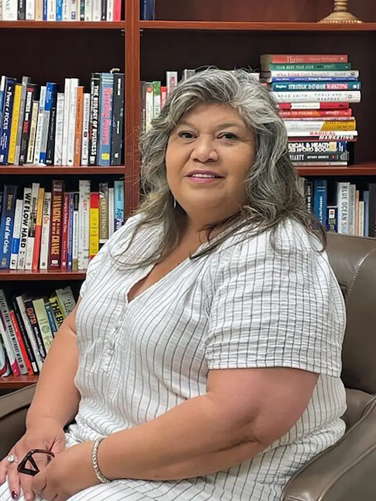 A woman with gray and brown hair sits in a brown chair, wearing a white striped shirt, in front of a bookshelf filled with books.
