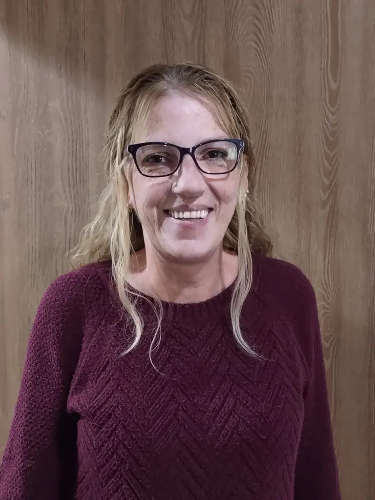 A woman with light brown hair and glasses, wearing a maroon sweater, stands in front of a wooden background and smiles at the camera. A woman with light brown hair and glasses, wearing a maroon sweater, stands in front of a wooden background and smiles at the camera.
