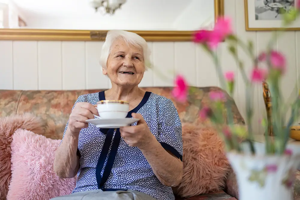 An elderly woman sits on a couch, smiling and holding a teacup and saucer, with pink cushions and a vase of pink flowers in the foreground.