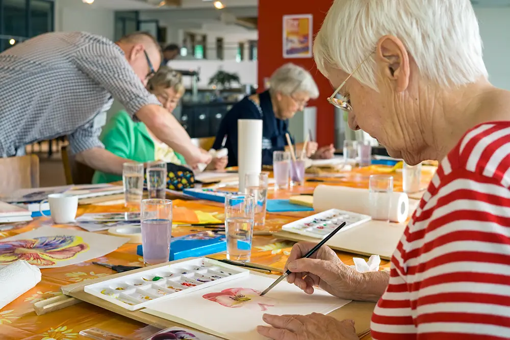 Several older adults sit at a table painting with watercolors in a well-lit room, while a man stands assisting one of the participants. Art supplies and glasses of water are on the table.