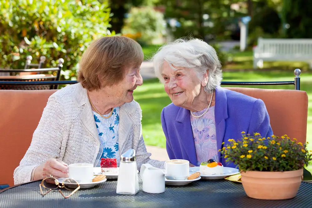 Two elderly women sit at an outdoor table, smiling and talking over coffee and pastries, with plants and greenery in the background.