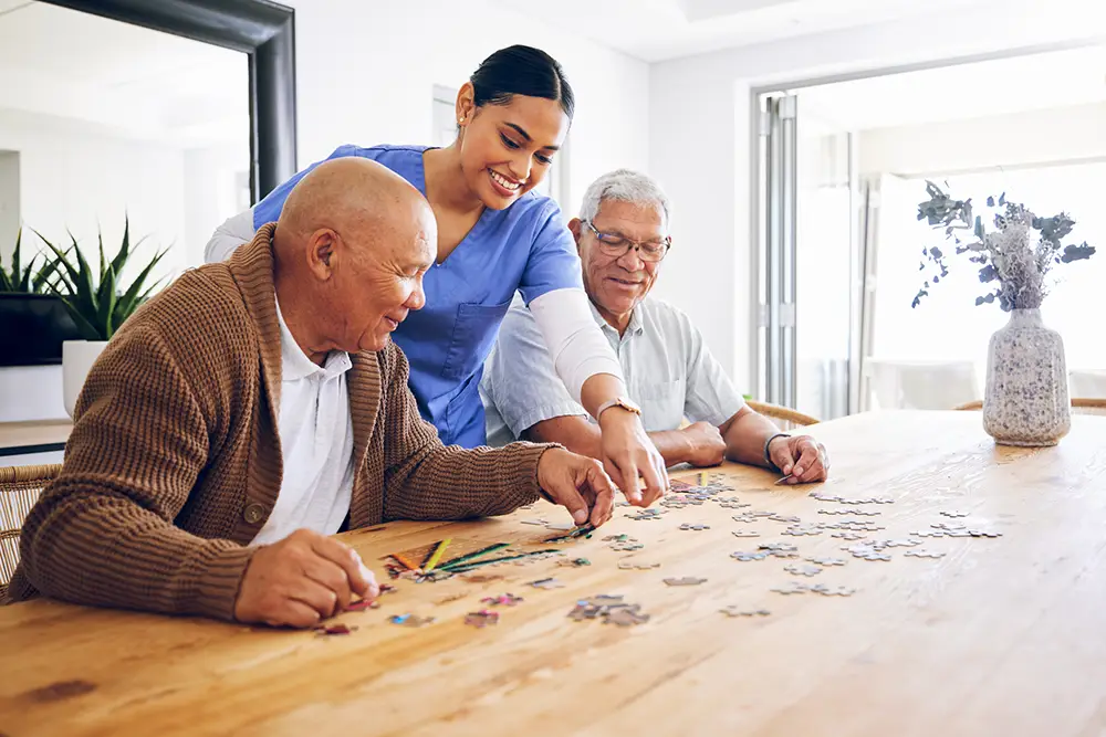 A caregiver assists two elderly men as they work on a jigsaw puzzle together at a wooden table in a bright, modern room.