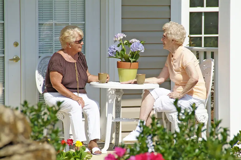Two elderly women sit at a small outdoor table with coffee mugs and a potted plant, talking outside a house on a sunny day.