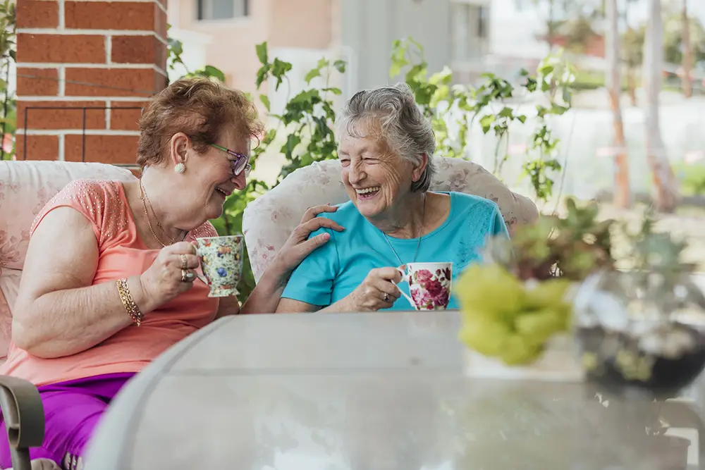 Two elderly women sit outside, smiling and laughing while holding floral mugs, with plants and a glass table in the foreground.