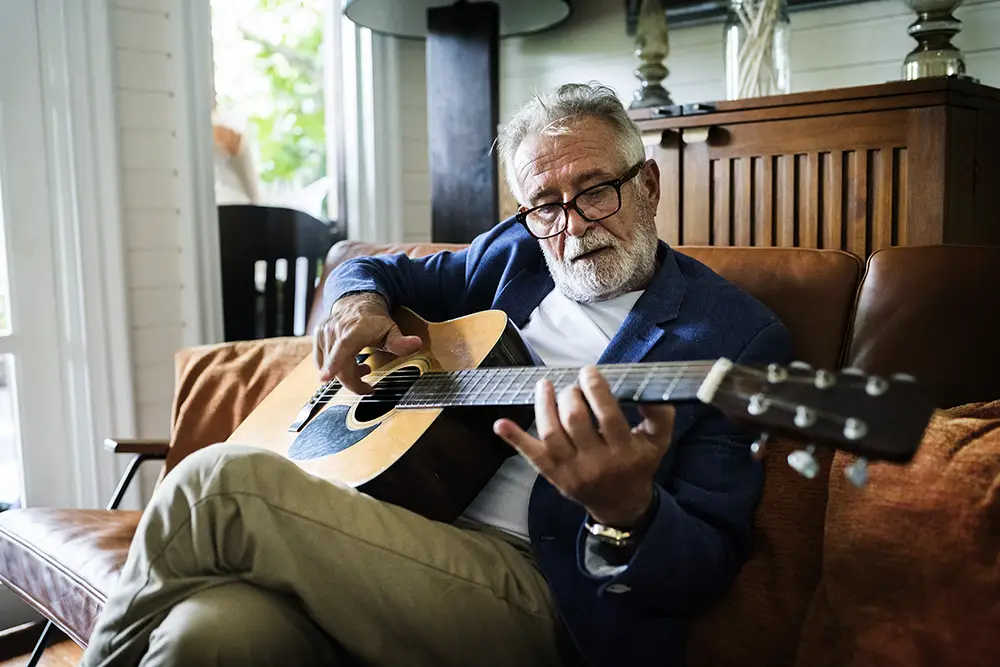 An older man with gray hair and glasses sits on a brown couch playing an acoustic guitar, wearing a blue blazer and light-colored pants.