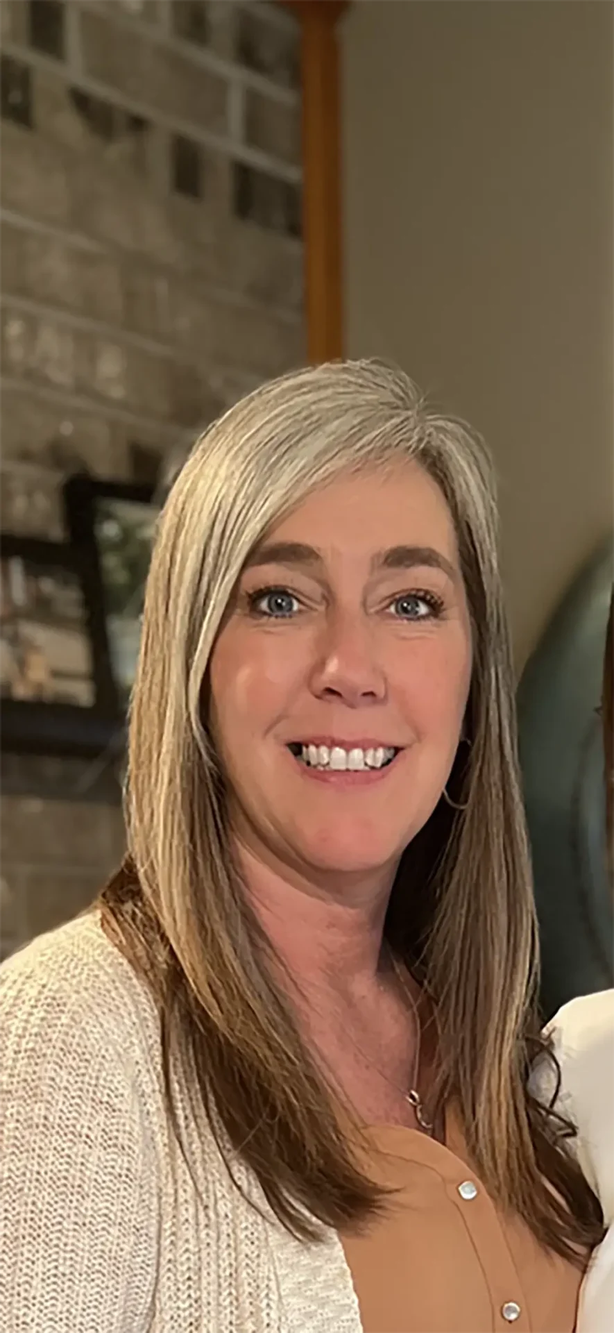 A woman with long, straight gray hair and blue eyes smiles at the camera indoors with a brick wall and framed photos in the background.