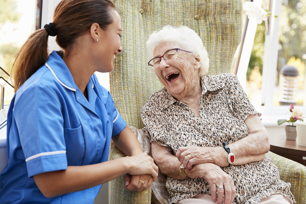 A caregiver in a blue uniform smiles and holds hands with an elderly woman, whose laughter brings joy as she sits in a chair—showing that laughter is truly good medicine.