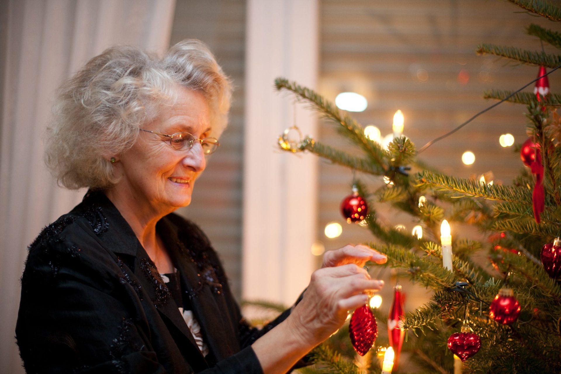 An older woman wearing glasses decorates a Christmas tree with red ornaments and lit candles in a warmly lit room, cherishing the holidays.