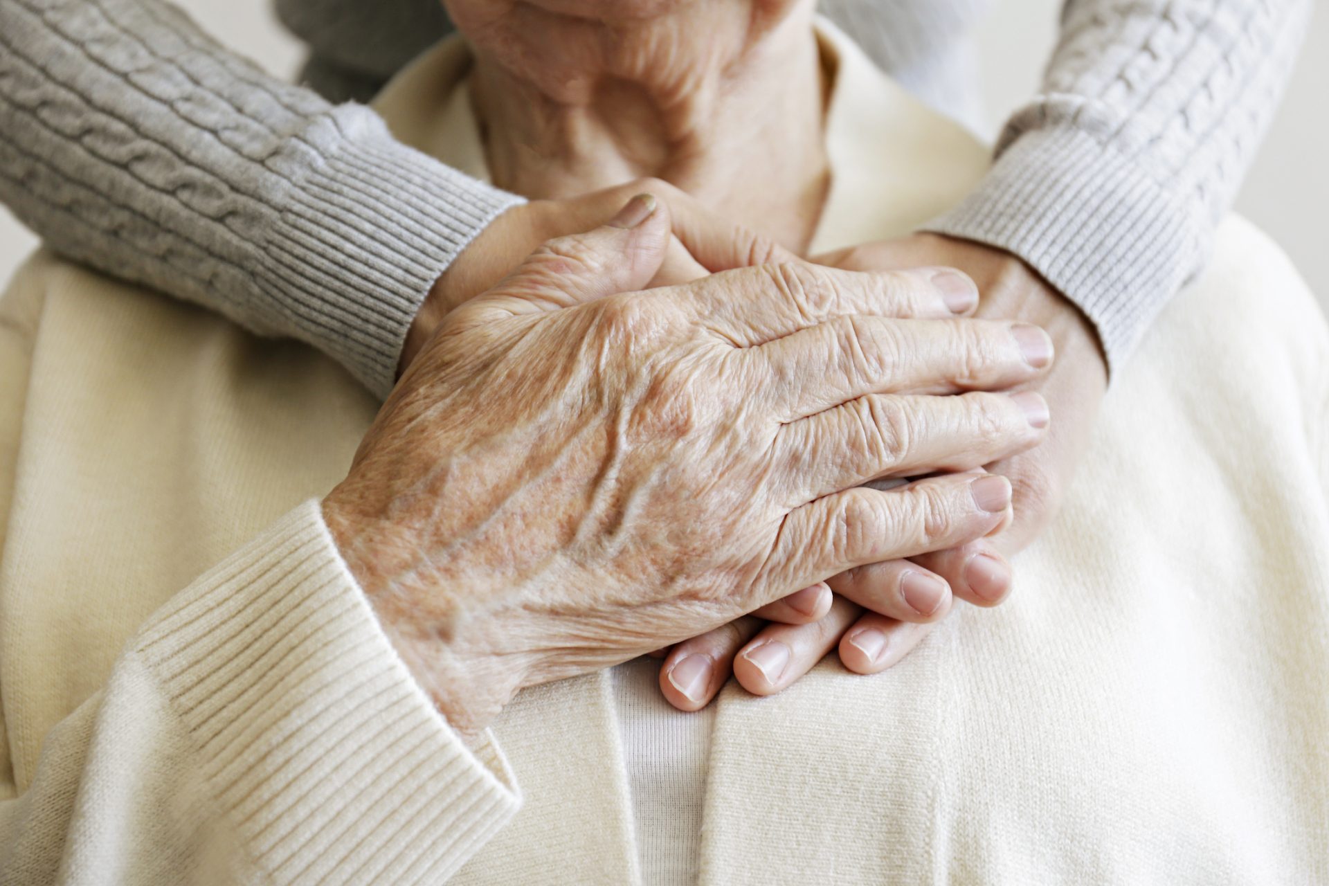An elderly person with their hands resting on younger hands, both wearing light-colored sweaters, the younger arms gently wrapped around the older person, beautifully illustrating the gift of aging and the importance of spiritual care.