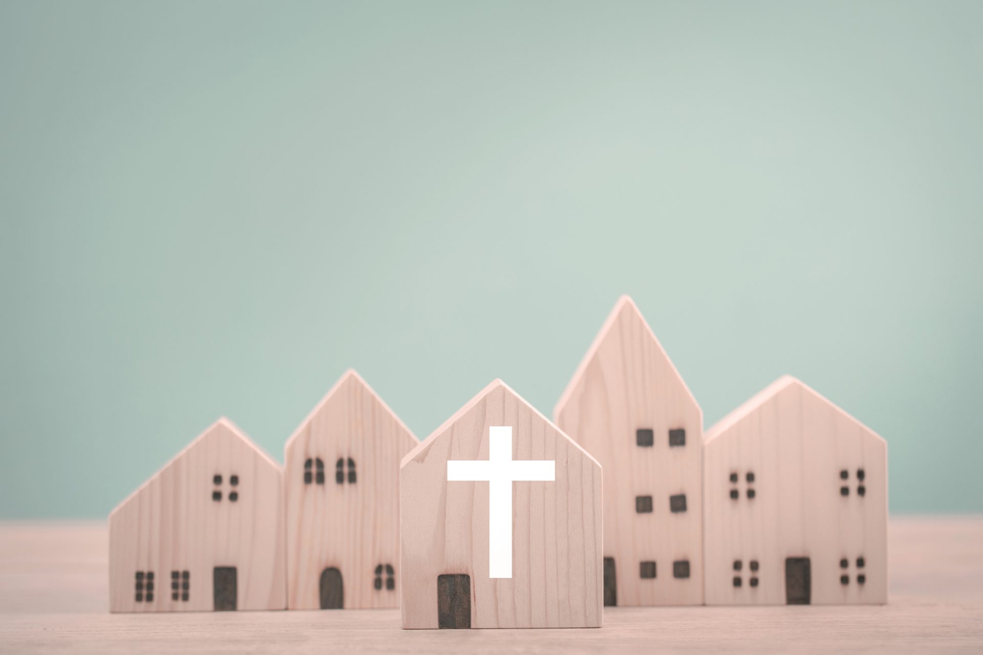 Several wooden toy houses are arranged side by side, with one house in the front displaying a white cross symbol, highlighting Spiritual Care. The background is plain light blue, emphasizing a sense of Great Connection within the community.