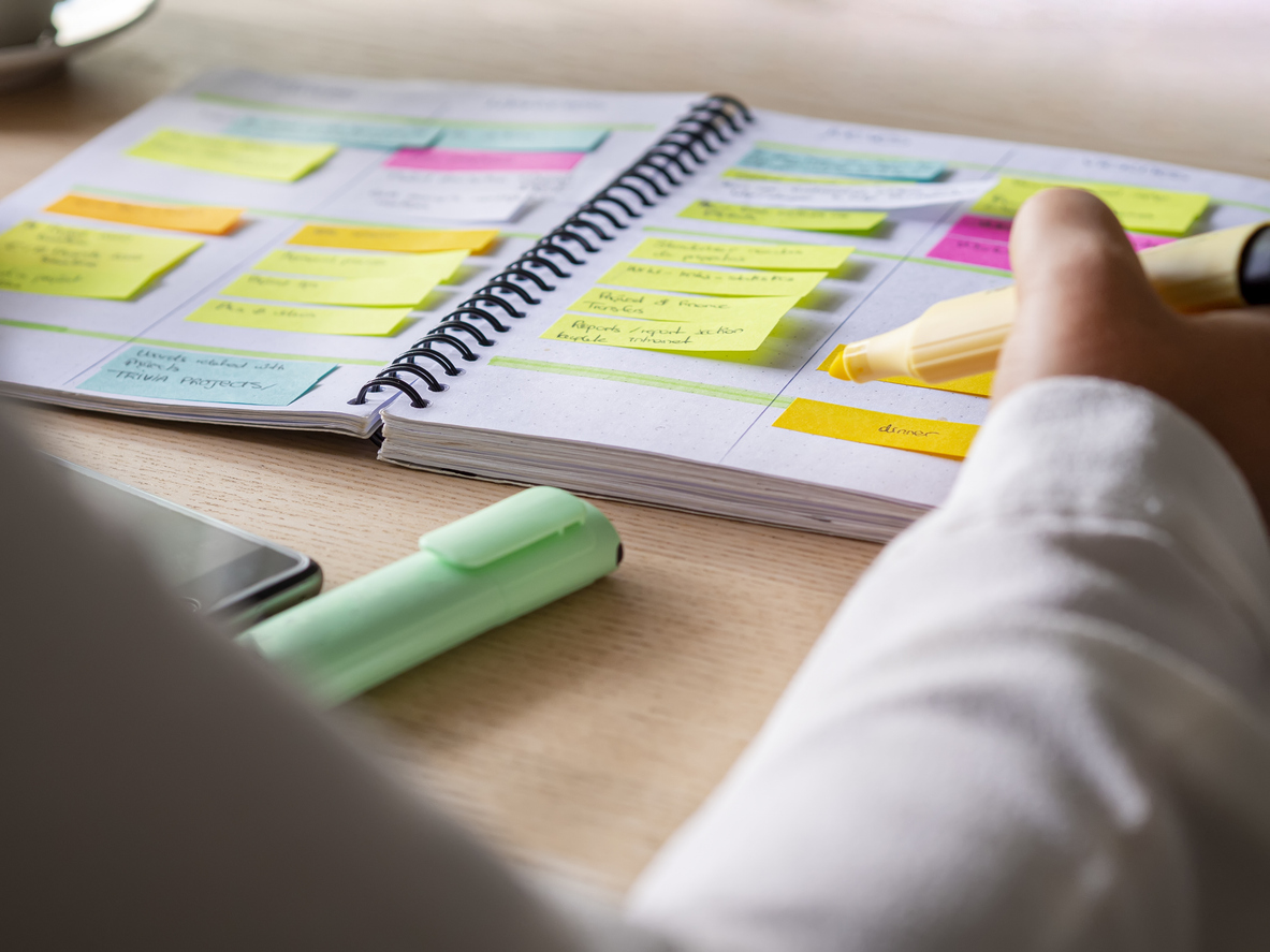 A person highlights notes in a spiral-bound planner filled with colorful sticky notes on a wooden desk, reflecting on Spiritual Care, with a highlighter and phone nearby.