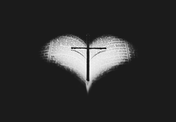 A cross casts a shadow in the shape of a heart onto the pages of an open Bible, symbolizing God's LOVE with focused lighting and a dark background.