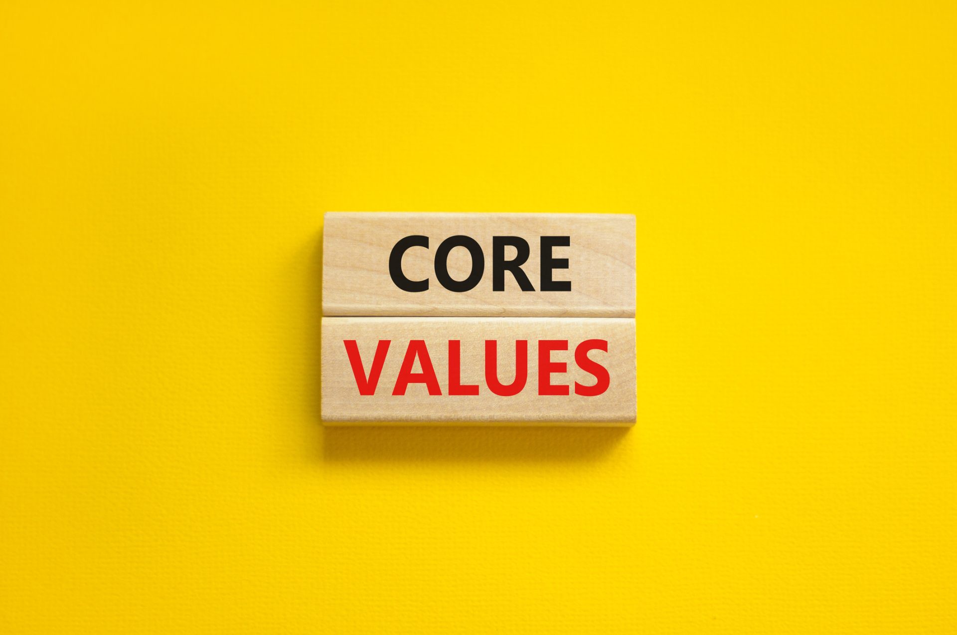 Two wooden blocks on a yellow background display "CORE VALUES"—"CORE" in black and "VALUES" in red—highlighting the importance of company values.