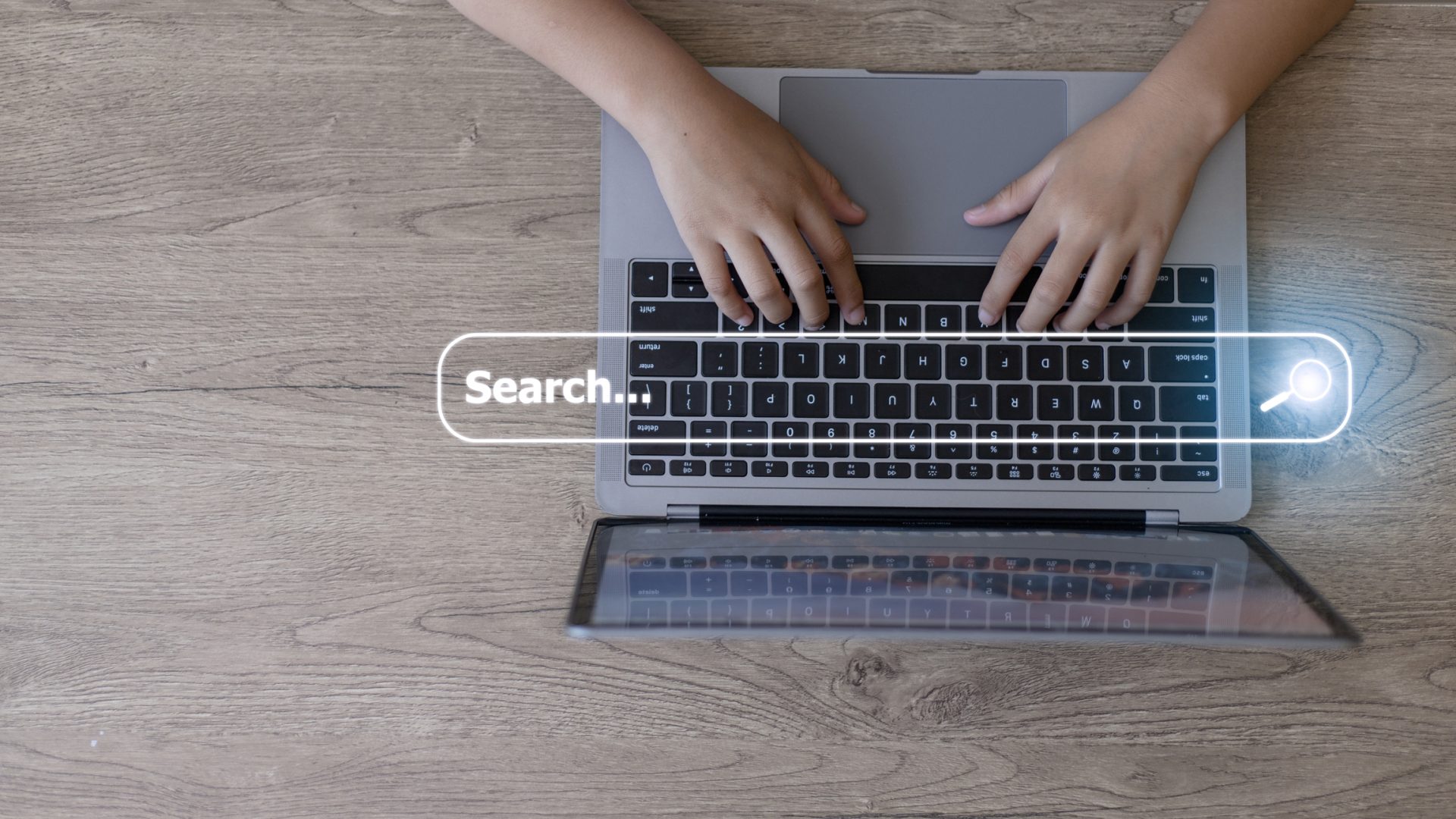 A person types on a laptop keyboard with a virtual search bar graphic overlaid above their hands on a wooden desk, searching for Senior Living Terminology.