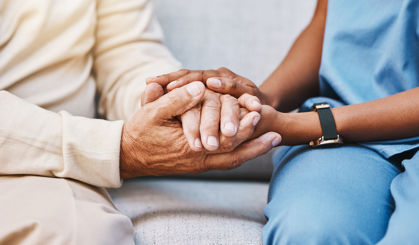 A person in medical scrubs holds the hands of an older person, both seated side by side on a light-colored couch, embodying how spiritual care can make a burden shared feel lighter—a theme featured in our November blog.