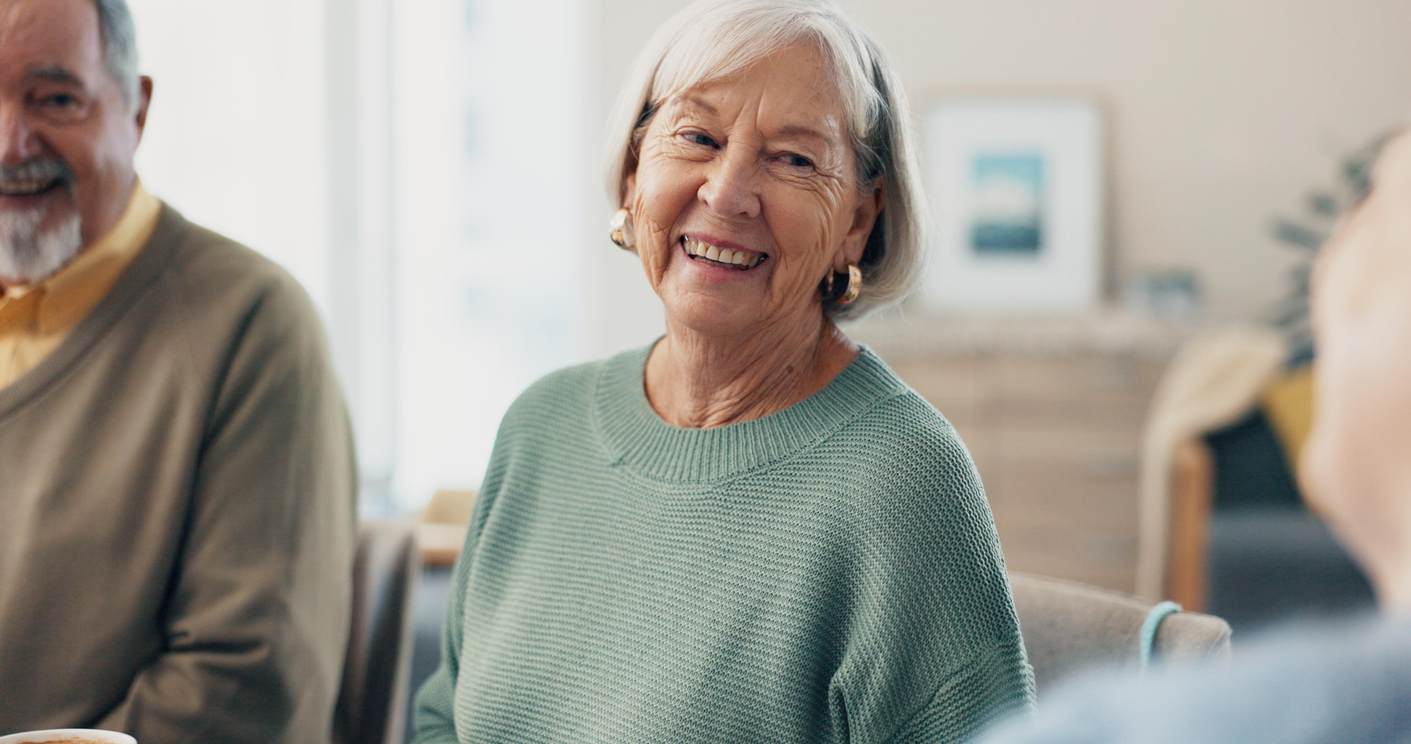 Older woman with gray hair and a light green sweater smiles while sitting indoors with others, sharing her success story and engaging in conversation.