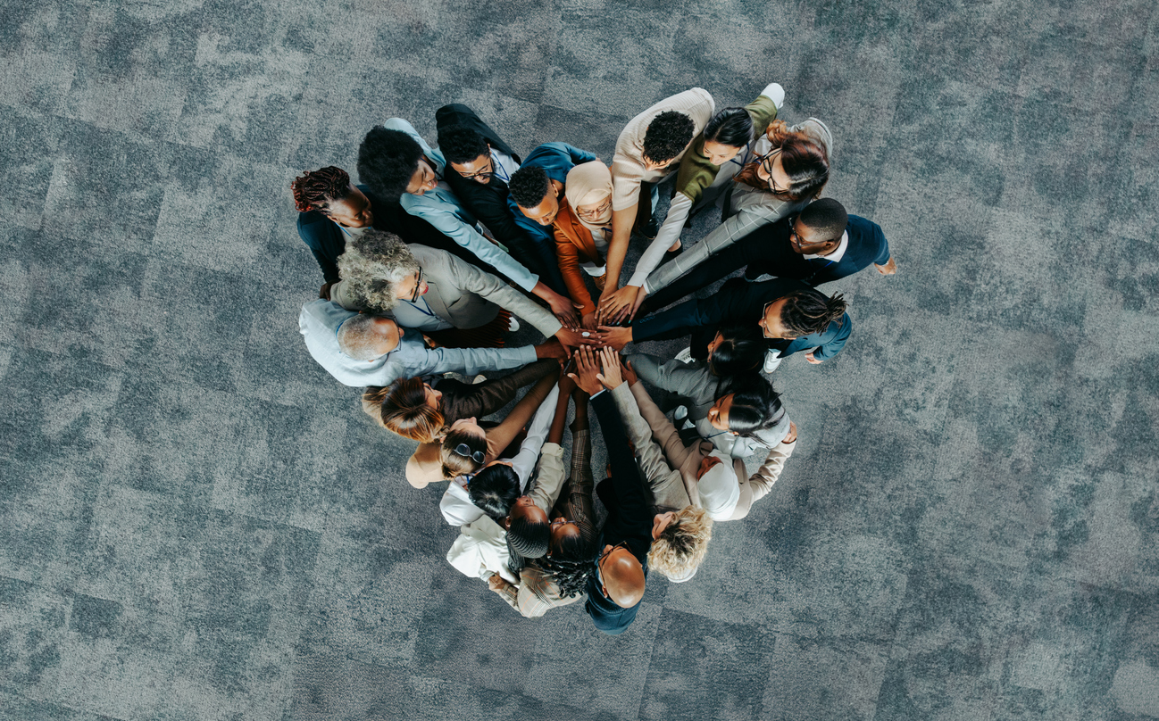 A group of people standing in a circle with their hands joined together in the center, symbolizing Living in Unity, viewed from above on a gray patterned floor.