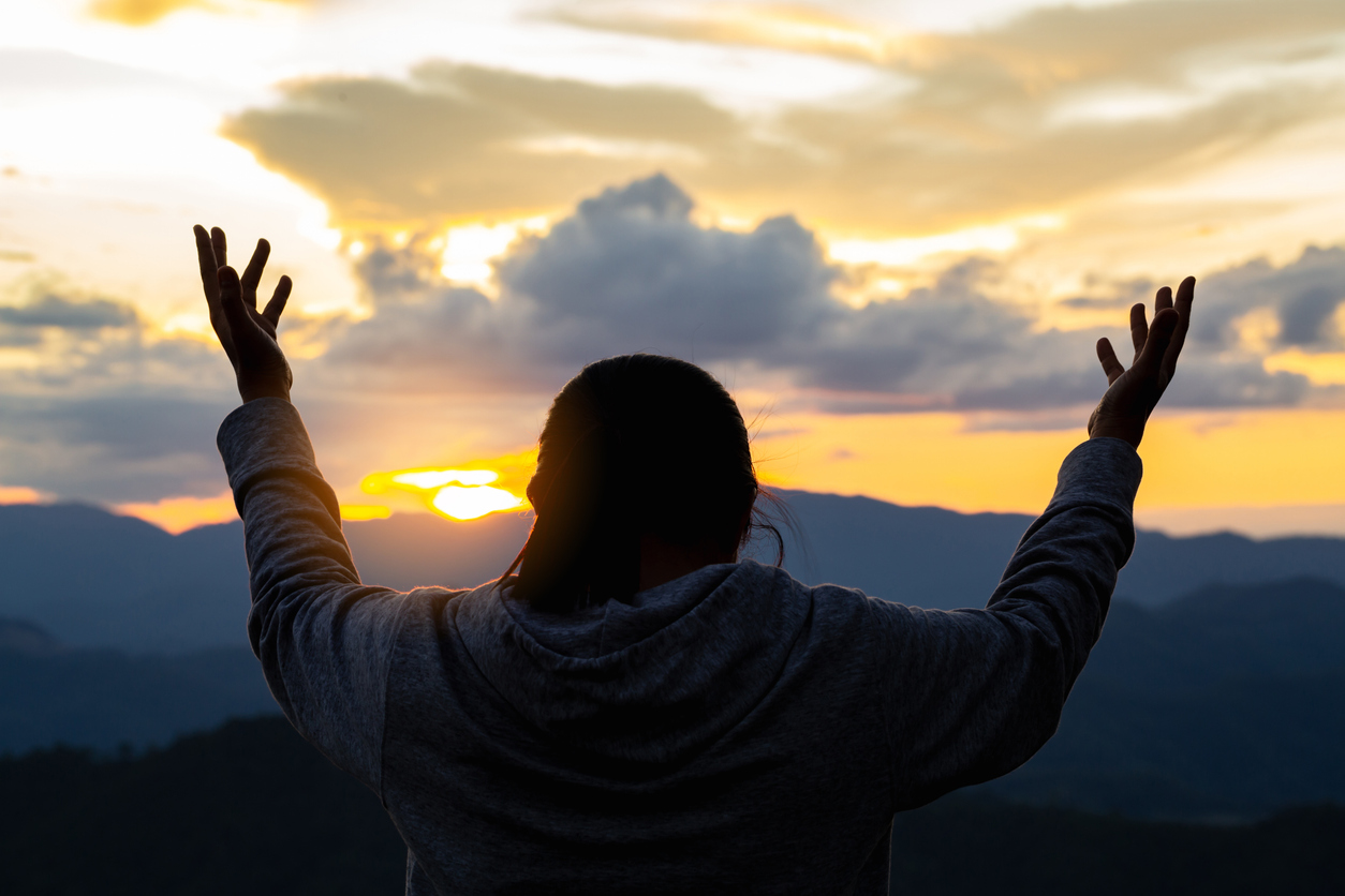 Silhouette of a person with raised arms facing a sunset over mountains, with clouds and a colorful sky—capturing the spirit of Pray Boldly and the essence of Spiritual Care in this peaceful October Blog moment.