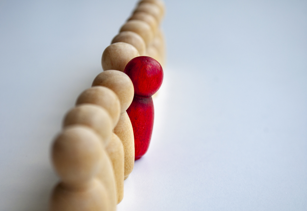 A row of light wooden figurines with one red figurine set apart in the center, standing out from the rest.