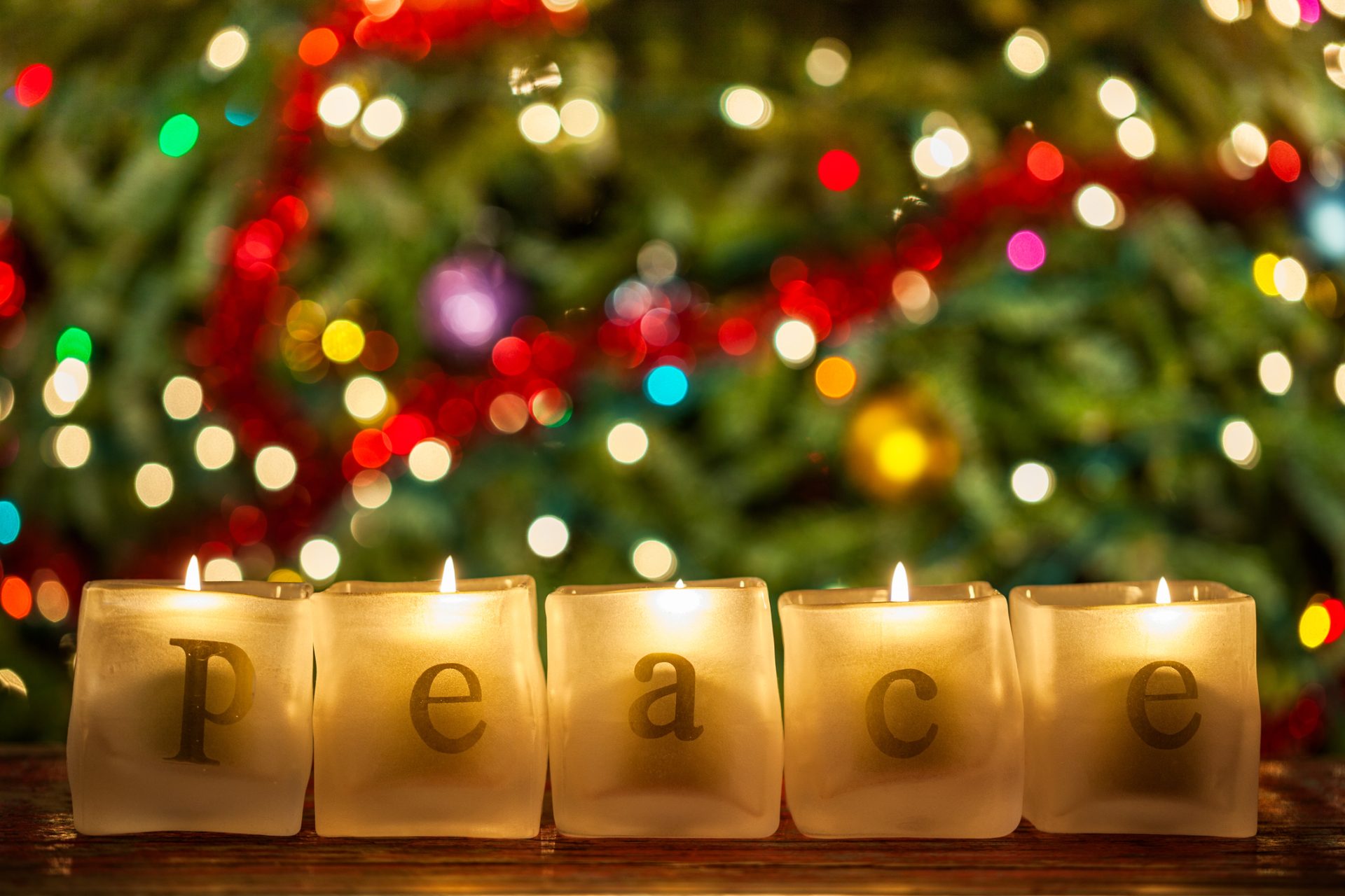 Five lit candles spelling "peace" sit in front of a blurred background with colorful Christmas lights and decorations, perfect for a December blog reflecting on spiritual care during the holiday season.