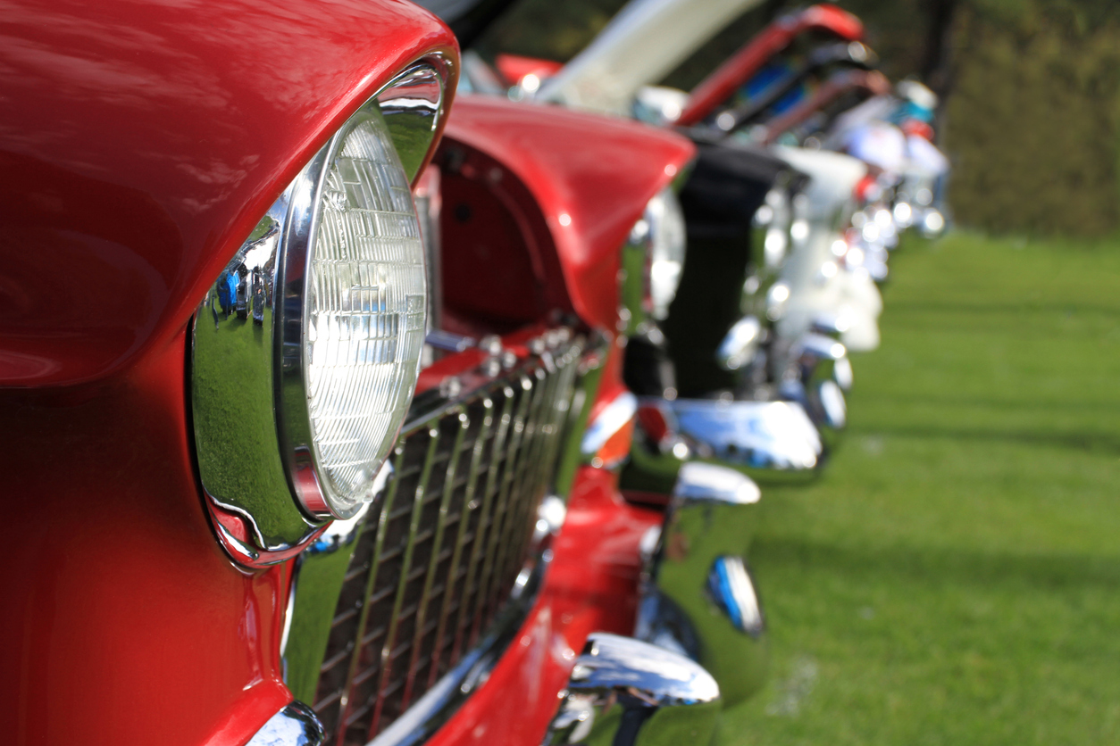Close-up of the front ends of several classic cars parked in a row on grass, with the focus on a red car in the foreground—a nostalgic scene that brings back memories of the good old days.