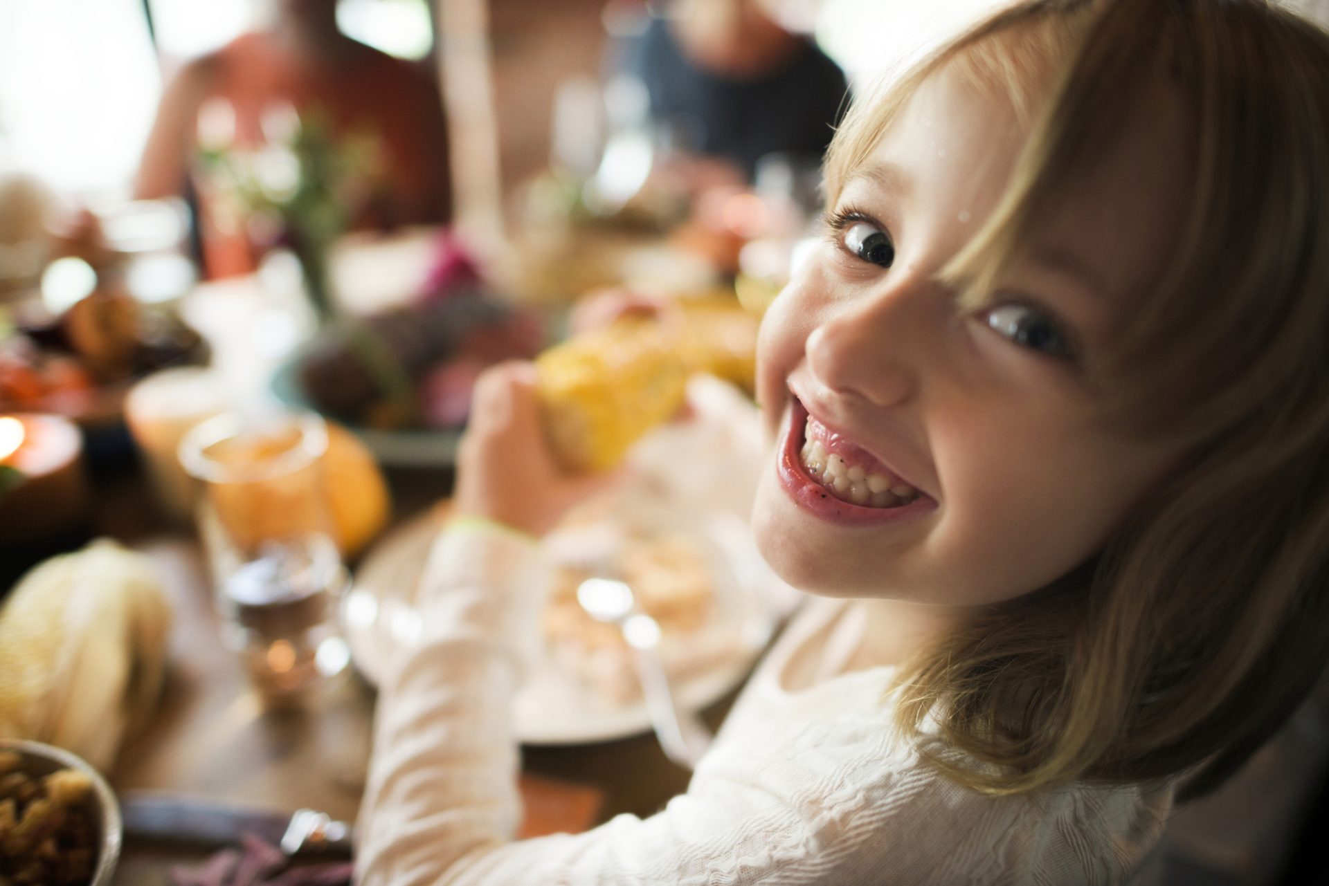 Smiling child holding and eating corn on the cob at a dining table with food and drinks in a warmly lit setting, radiating grace and joy reminiscent of the kindness Bob Goff writes about.