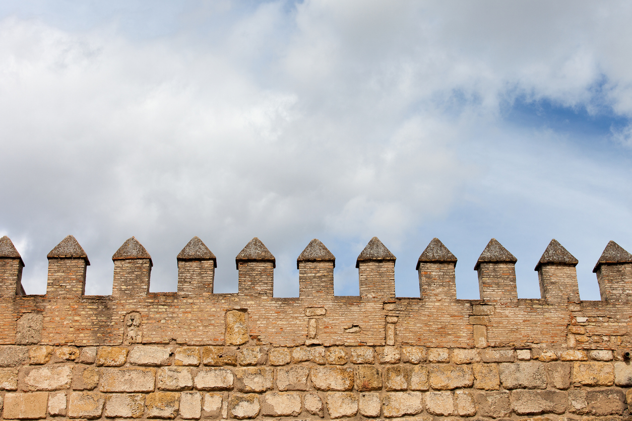 Stone castle wall with rectangular merlons, standing as a steadfast fortress against a cloudy sky, offering a sense of refuge.