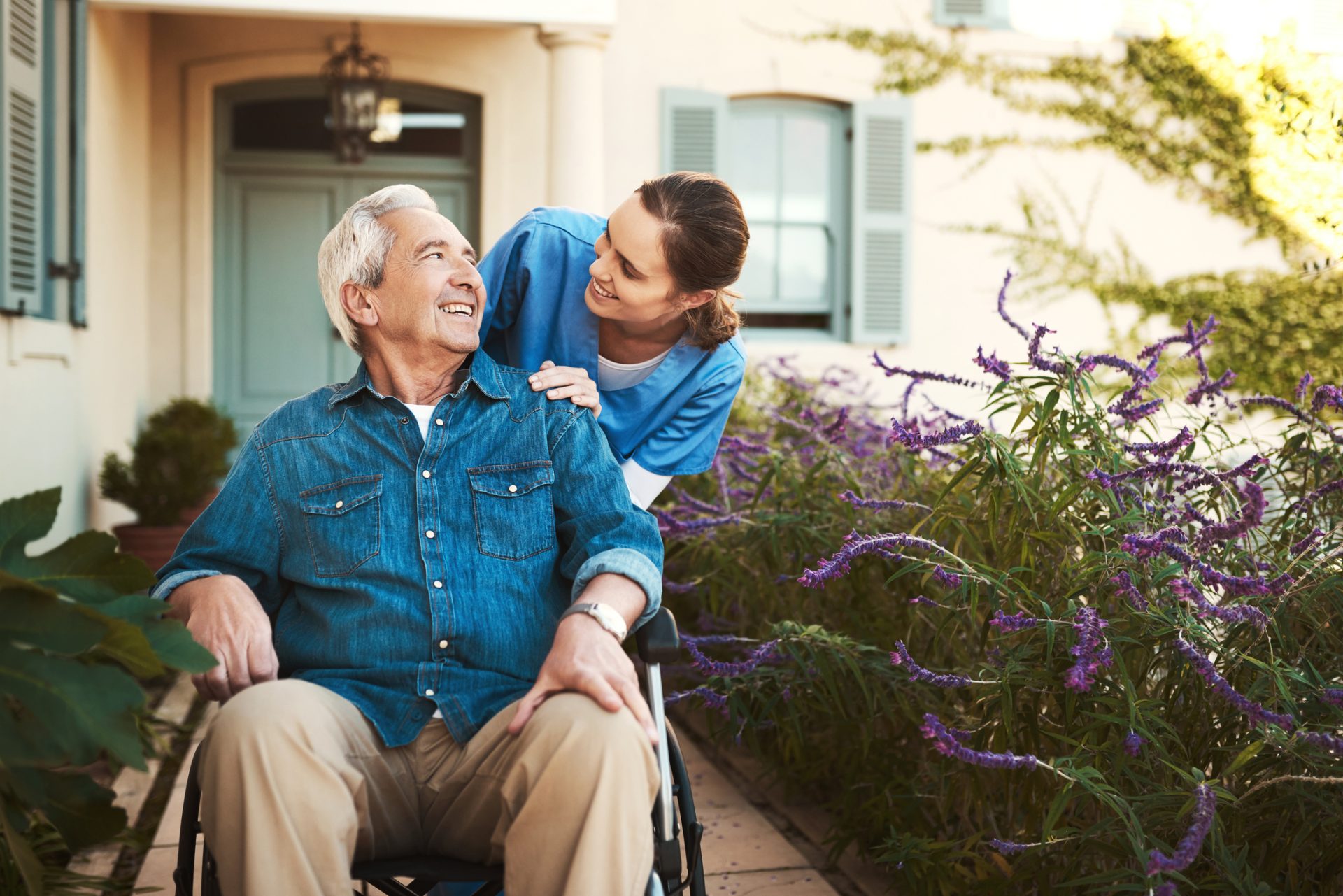 An older man in a wheelchair sits outside by purple flowers while a caregiver from an assisted living community stands behind him, both smiling at each other.