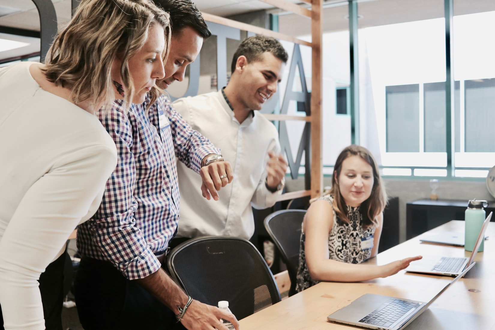 Four people in business attire gather around a desk, looking at a laptop screen and discussing an employee recognition program in an office setting.