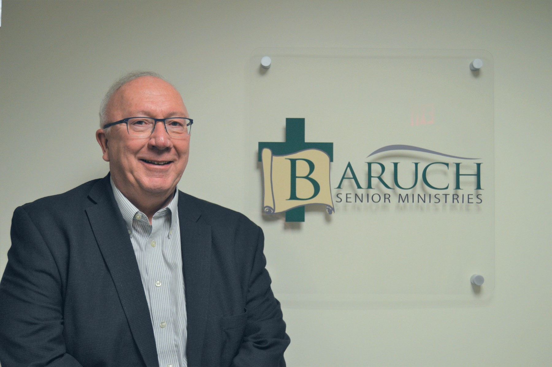 A man in a suit stands beside a wall sign that reads "Baruch Senior Ministries," celebrating the organization's 25th Anniversary.