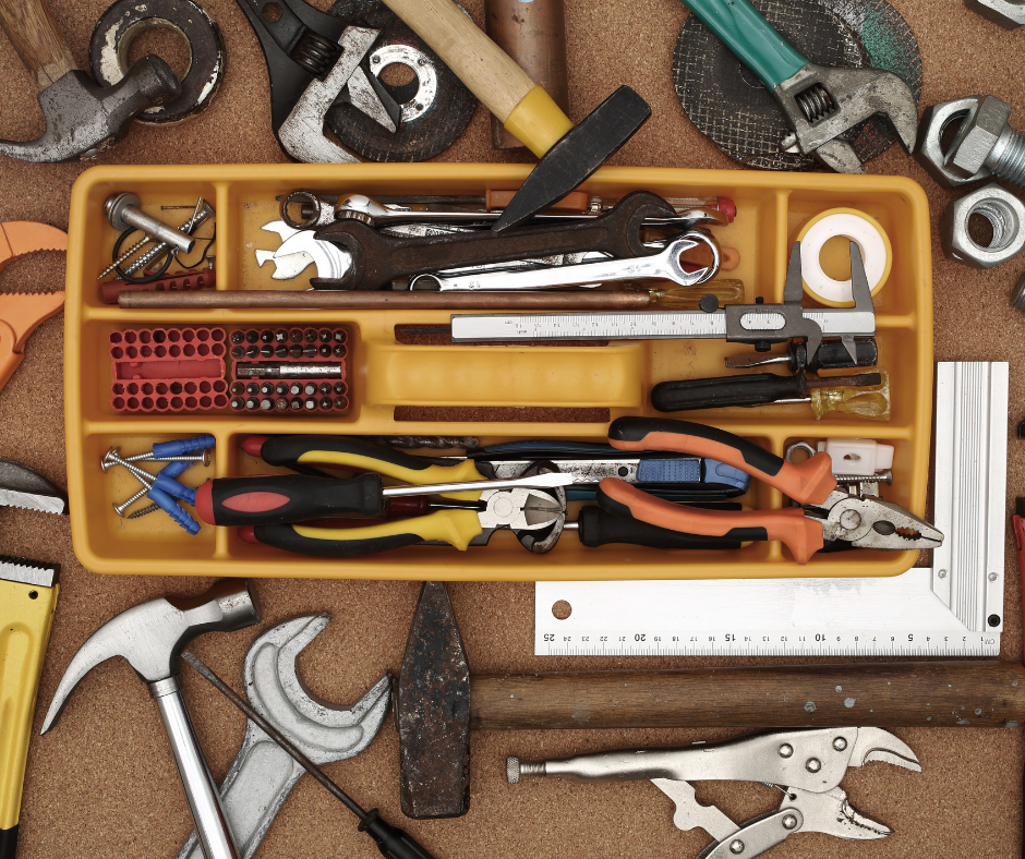 Top-down view of a yellow toolbox—the perfect addition to any Caregiver’s Toolbox—containing various hand tools, with more tools, including hammers and pliers, spread out on the work surface around it.
