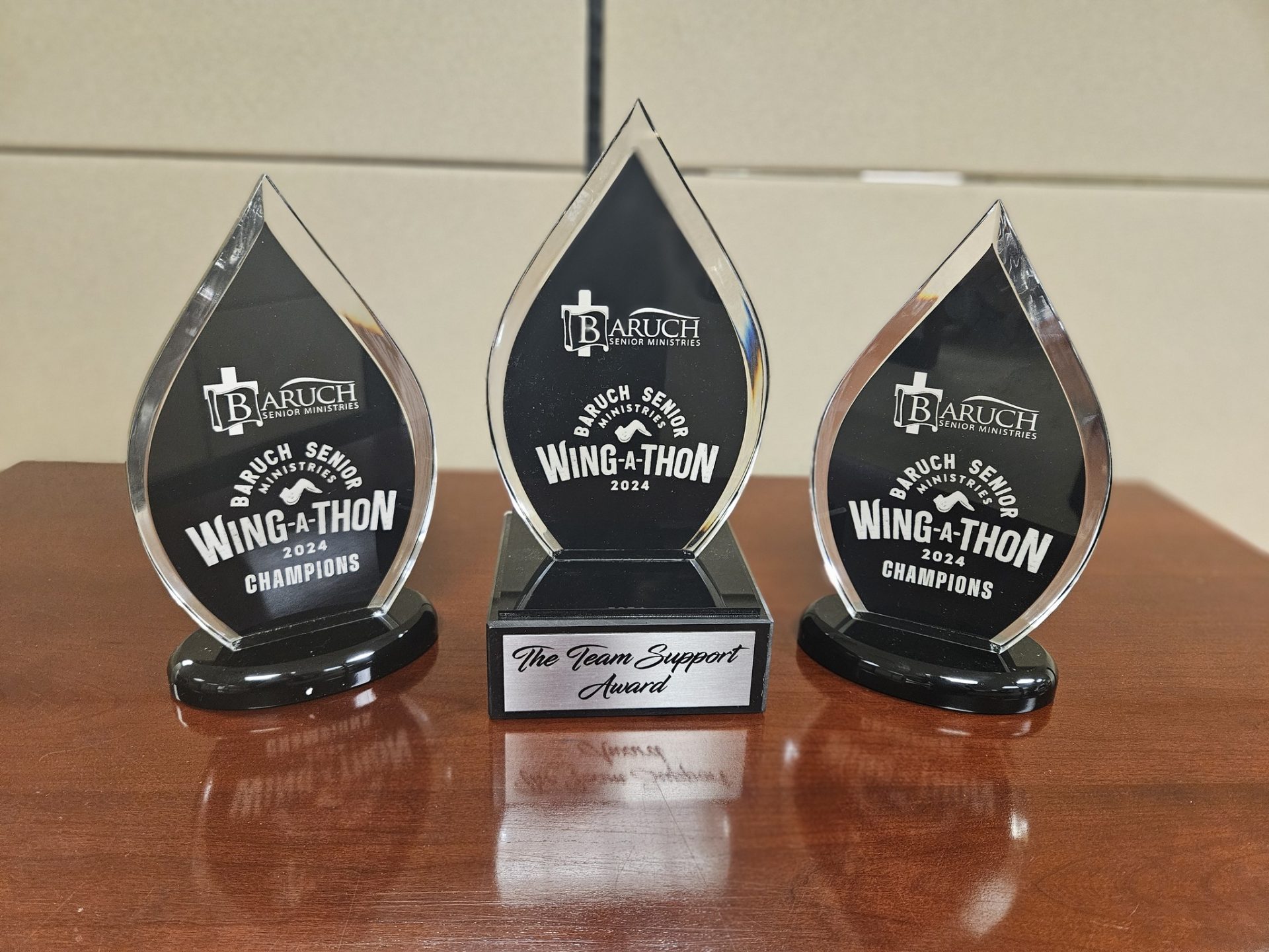 Three Baruch Senior Wing-A-Thon 2024 Champions trophies and a Team Support Award from the wing competition are displayed on a wooden surface, celebrating impressive results.