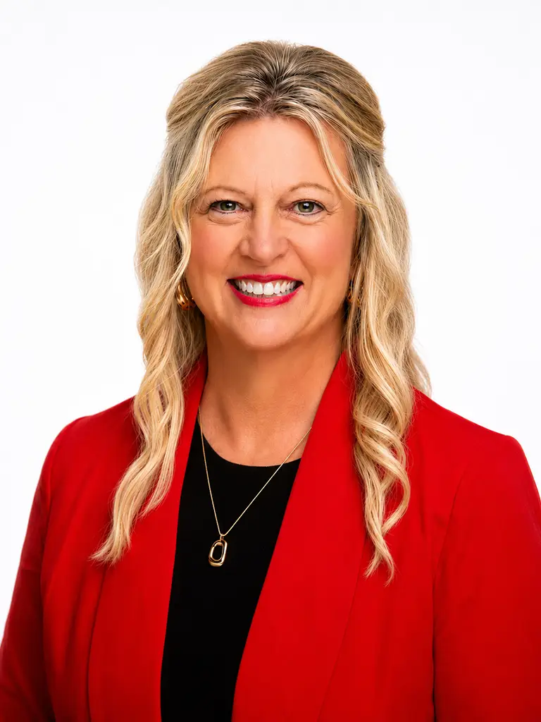 Smiling woman with long blonde hair wearing a red blazer, black top, and gold necklace, posing against a plain white background.